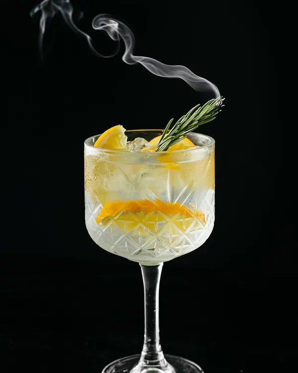 Copa balón con hielo, rodaja de naranja y una rama de romero, con un hilo de humo saliendo del vaso.