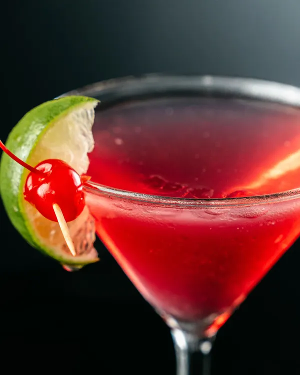 Cóctel rojo en copa de martini, con una rodaja de limón y una cereza en el borde.