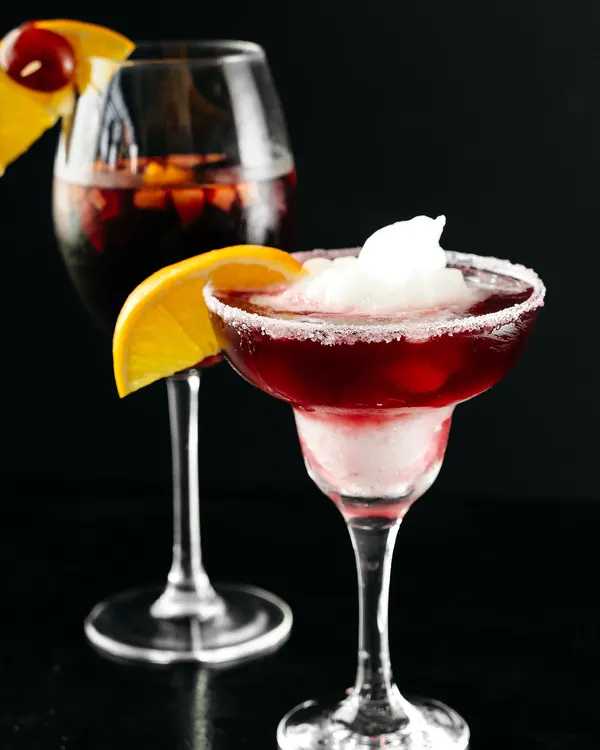 Copa de vino tinto con fruta y una rodaja de naranja, junto a una margarita roja con borde de azúcar.