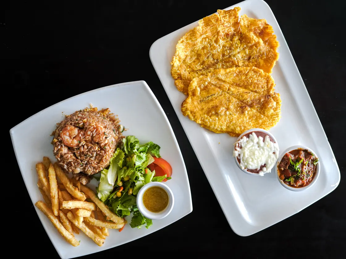 Dos platos sobre fondo negro: arroz con camarones, papas fritas y ensalada; y patacones con dos acompañamientos.