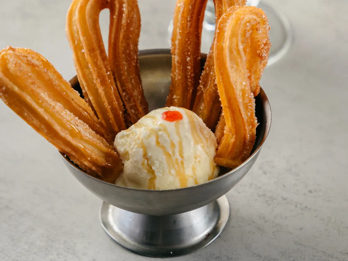 Churros en una copa metálica con una bola de helado, y un café con espuma detrás.