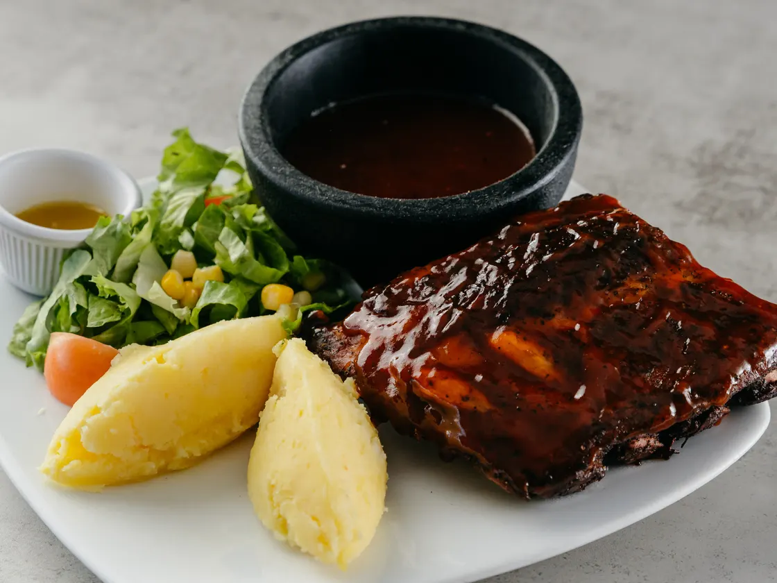 Costillas glaseadas en salsa oscura, con puré de papa, ensalada y un tazón negro con salsa.