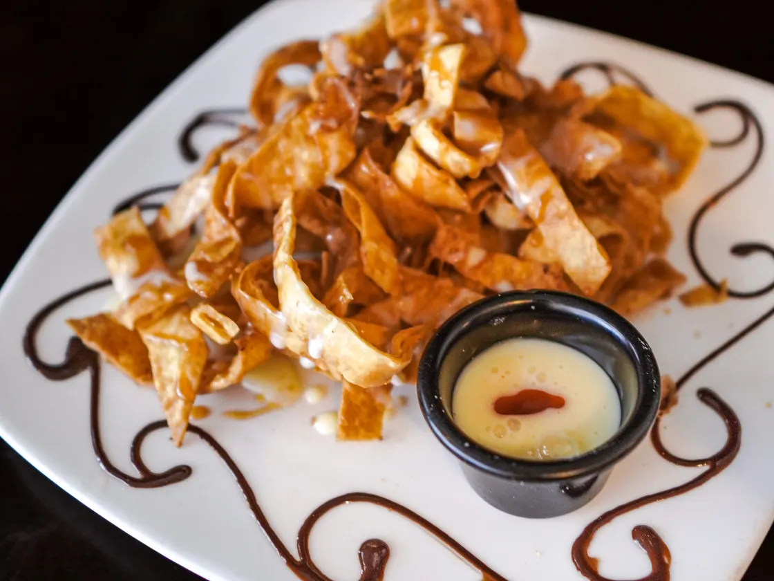 Tiras fritas y crujientes bañadas en caramelo, en un plato decorado con chocolate y con una salsa cremosa aparte.
