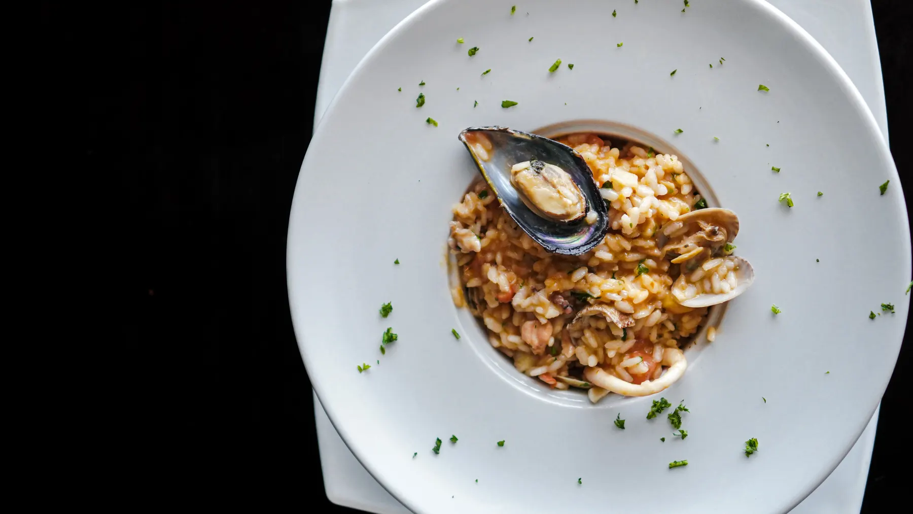 Arroz con mariscos visto desde arriba en un plato hondo blanco, con mejillón, almejas y calamar, sobre fondo negro.