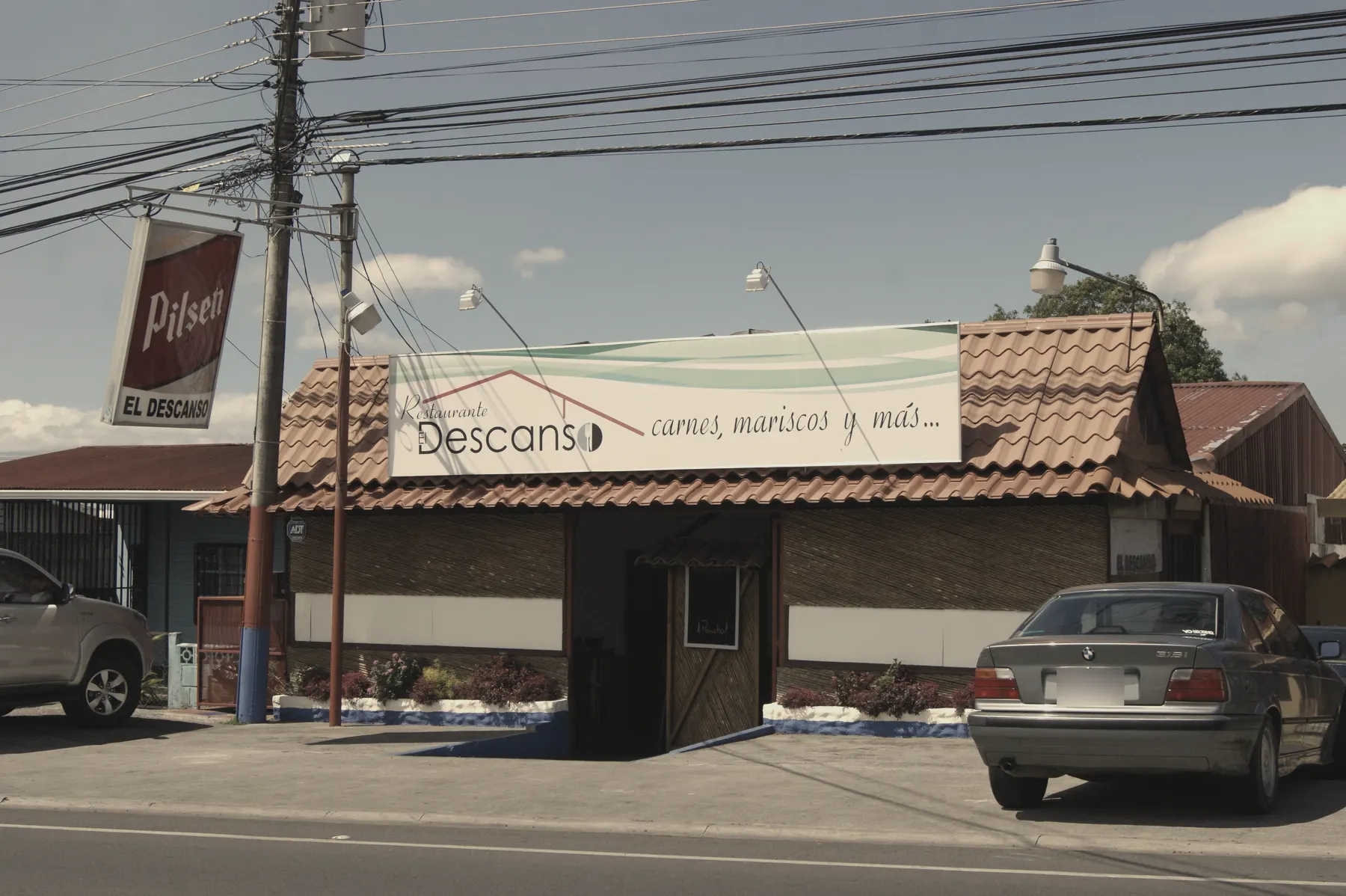 La fachada de El Descanso en otra época: techo de teja, paredes de caña y el rótulo «Restaurante El Descanso, carnes, mariscos y más…». A la izquierda, un rótulo de Pilsen con el nombre del lugar.